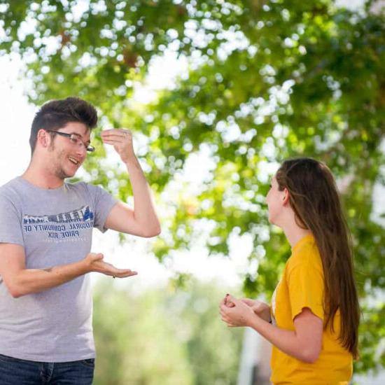 Student signing to another while under a tree.