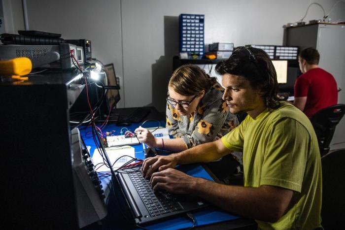 Two individuals are working together at a desk equipped with electronic components and a laptop. The man uses the laptop, while the woman interacts with electronic devices and wires while looking at the computer screen. Another person is in the background focusing on their computer. The room is dimly lit.