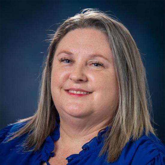 A smiling woman with shoulder length blonde straight hair, wearing a navy blue shirt, is set against a dark blue background.